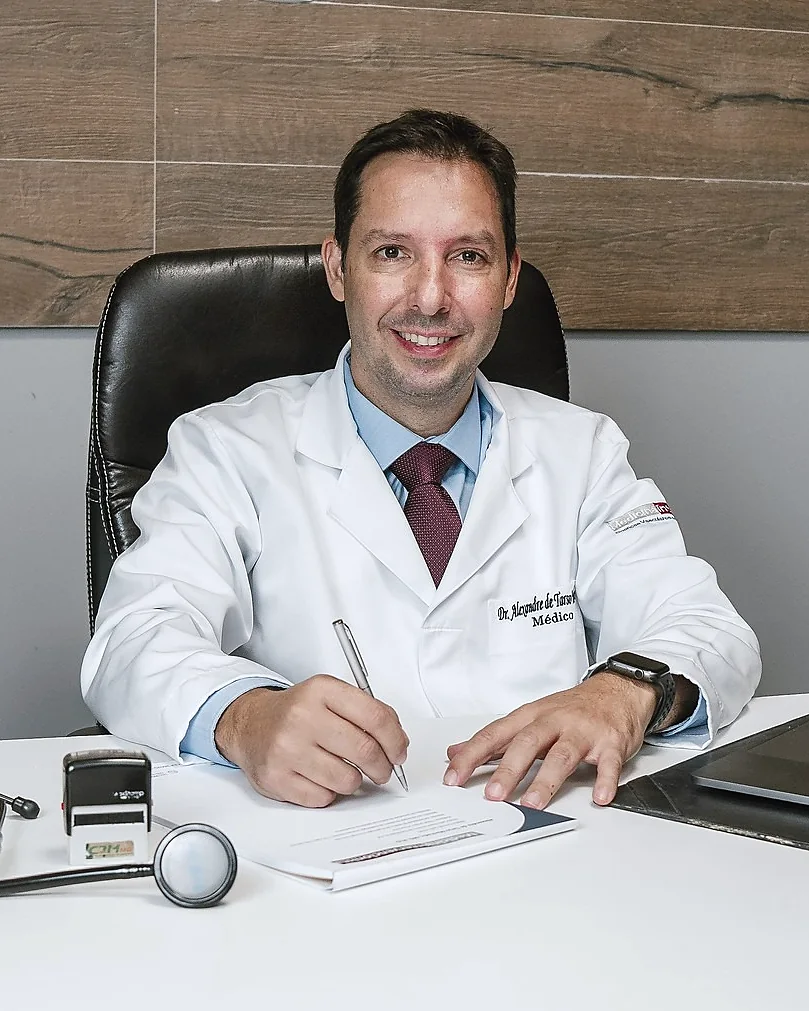 Dr. Alexandre de jaleco, sentado à mesa do consultório, escrevendo e sorrindo