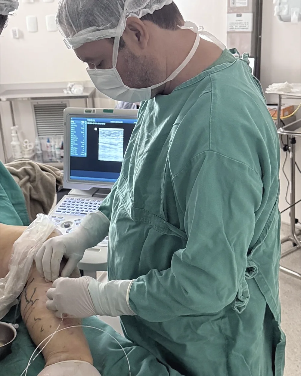 Dr. Alexandre paramentado fazendo uma punção na perna guiada pelo ultrassom, com a imagem no monitor ao fundo