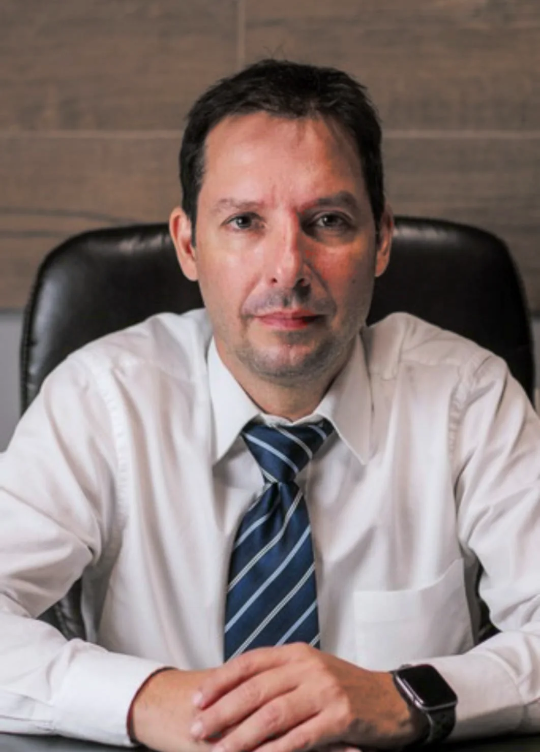Dr. Alexandre de Tarso Machado, de camisa branca e gravata azul listrada, sentado à mesa com as mãos cruzadas, em retrato de estúdio